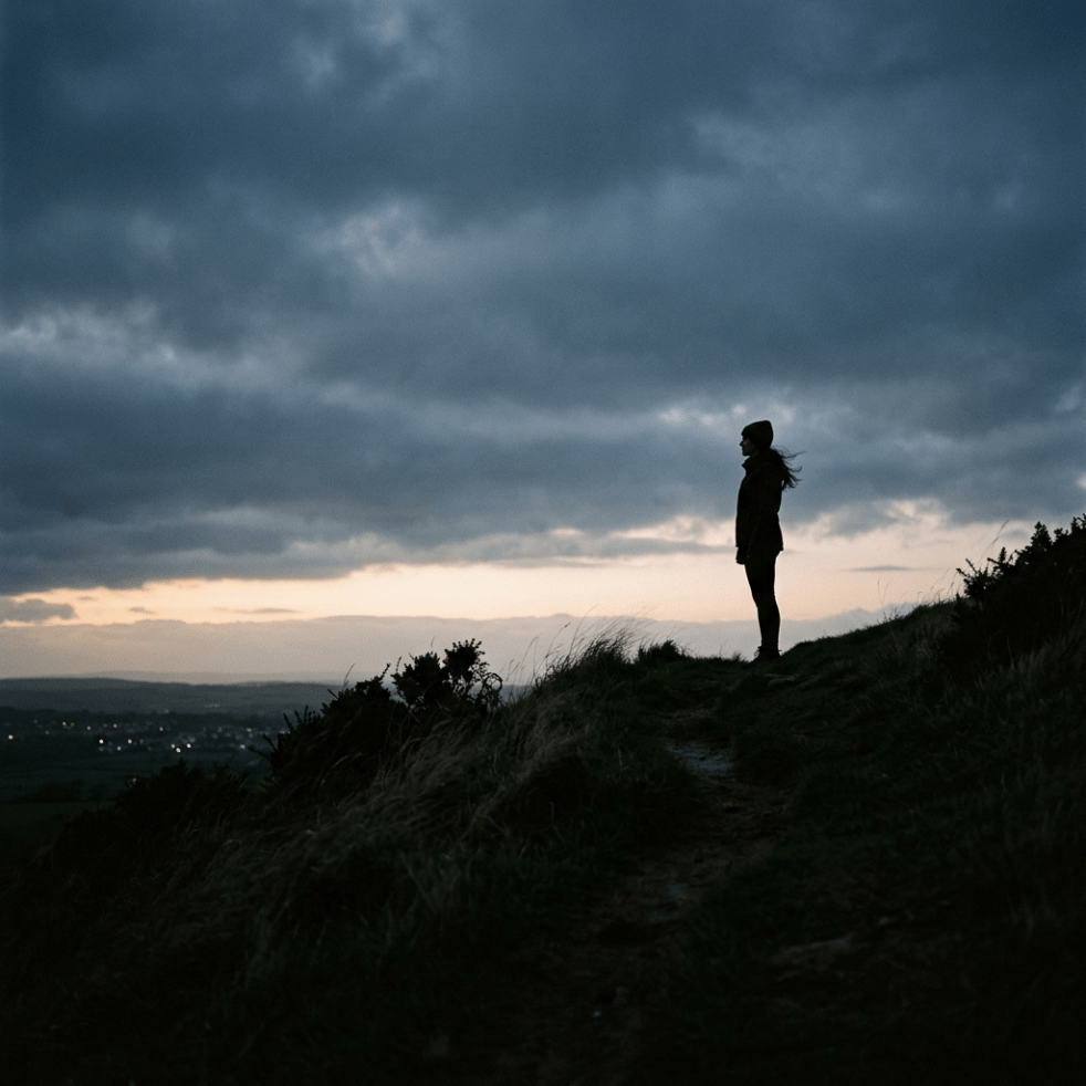Silhouette of a person standing on a grassy hill at dusk with cloudy sky